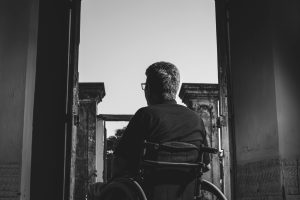 Black and white photo of a man in a wheelchair looking outside through an open doorway, evoking solitude.